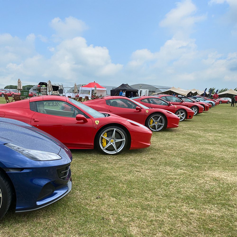 A row of red Ferrari sports cars and one blue Ferrari are parked on a grassy field under a partly cloudy sky, with event tents and people visible in the background.
