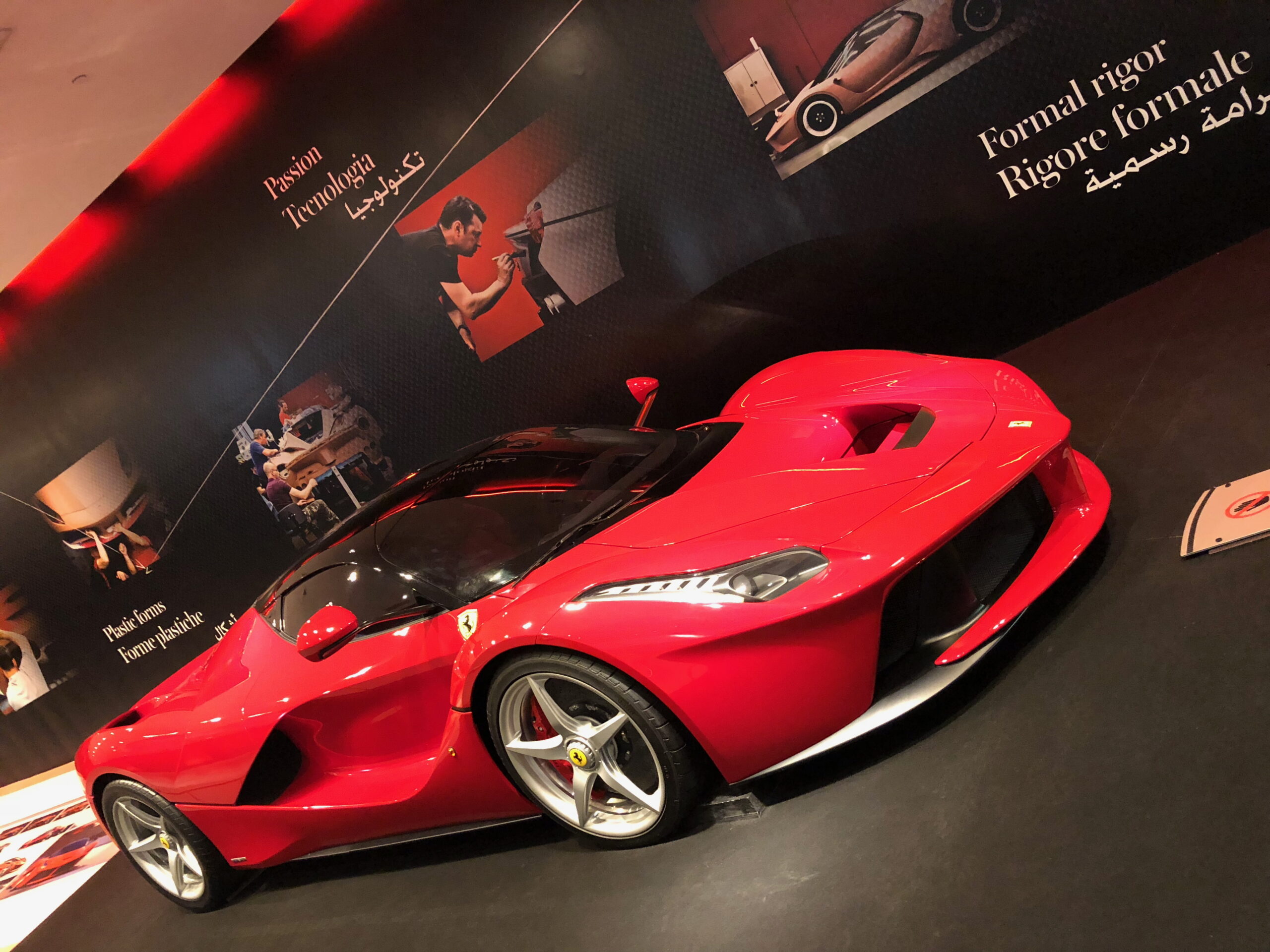 A red Ferrari sports car is displayed indoors on a black floor at Ferrari World Abu Dhabi, with informational posters and images about Ferrari’s craftsmanship and design in English, Italian, and Arabic on the wall behind it.