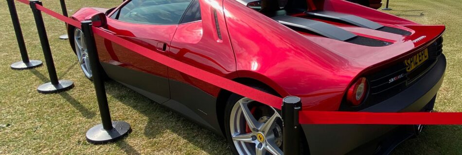 A shiny red sports car, inspired by Eric Claptons Ferrari SP12EC, is parked on grass behind red and black barrier posts, with another similar car nearby. The scene unfolds in an open field under a partly cloudy sky.