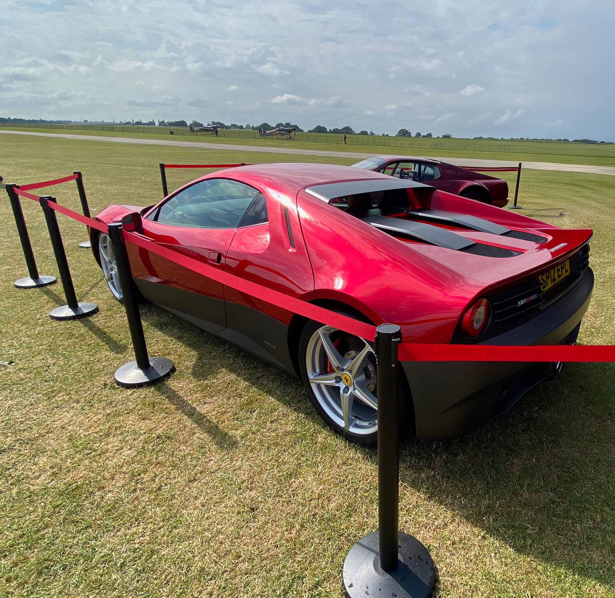 A shiny red sports car, inspired by Eric Claptons Ferrari SP12EC, is parked on grass behind red and black barrier posts, with another similar car nearby. The scene unfolds in an open field under a partly cloudy sky.